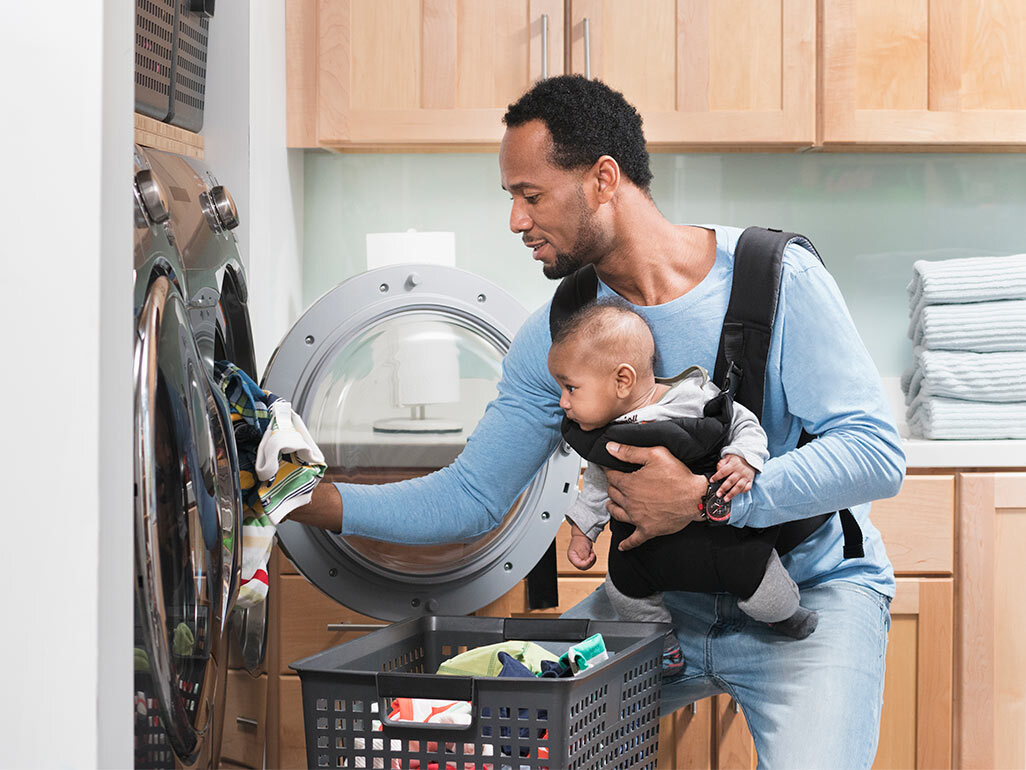 A man wearing a baby while loading clothes into a washing machine