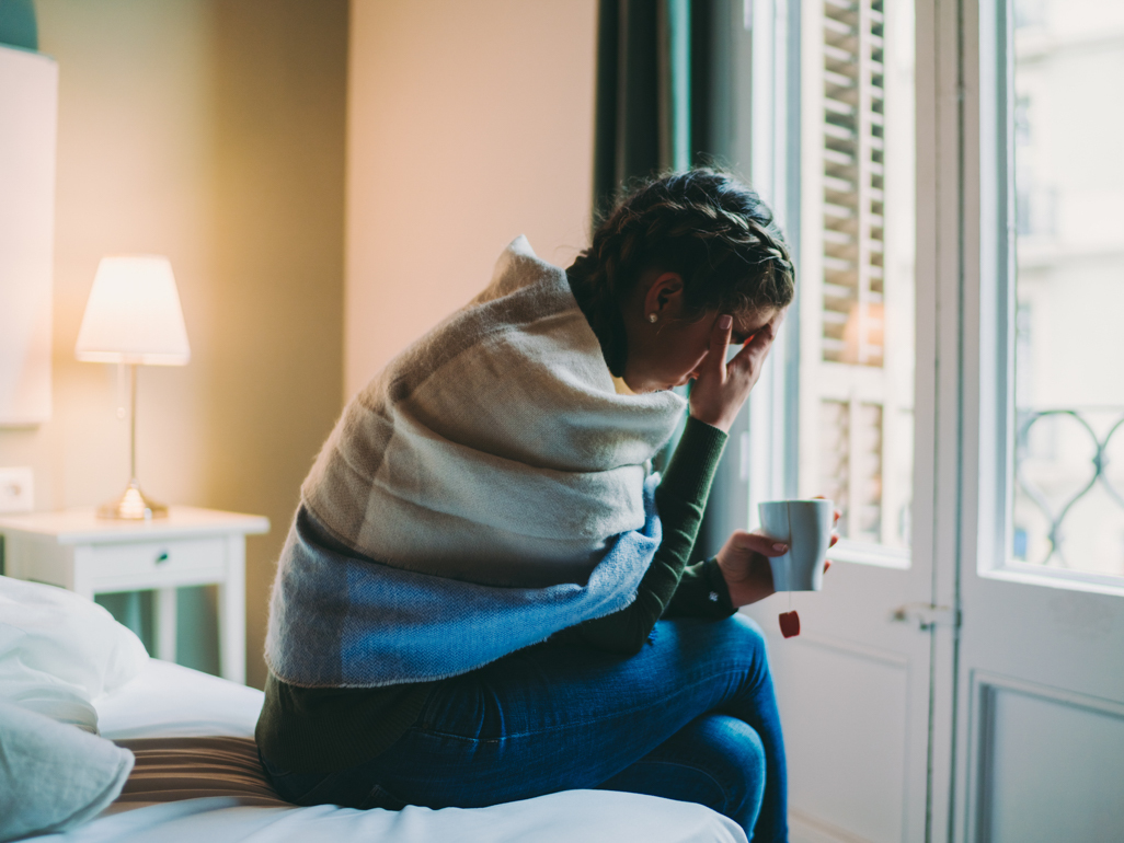 woman suffering from nausea sitting on bed hunched over drinking tea