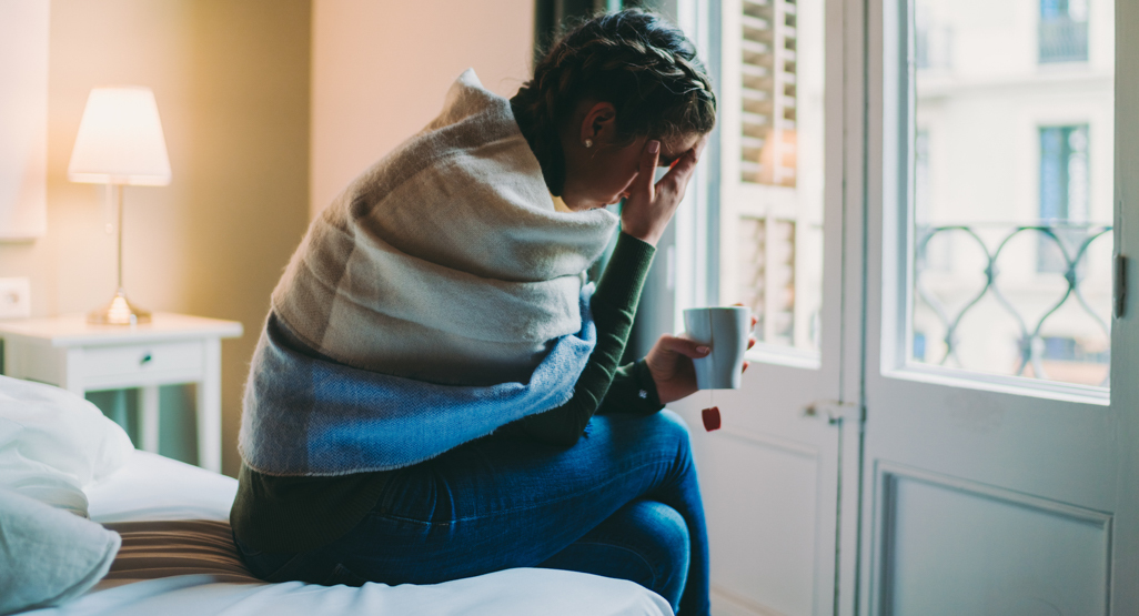 woman suffering from nausea sitting on bed hunched over drinking tea