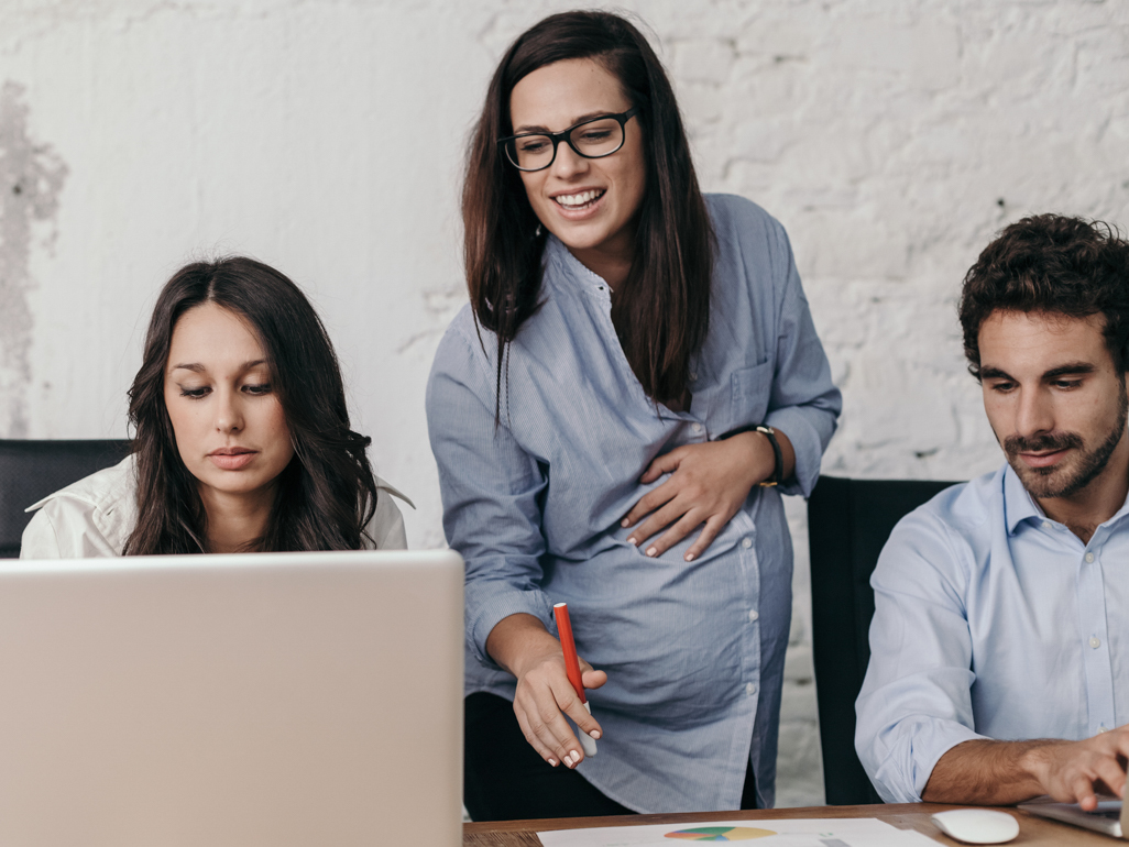 A pregnant woman at work looking over her coworkers' shoulder. 